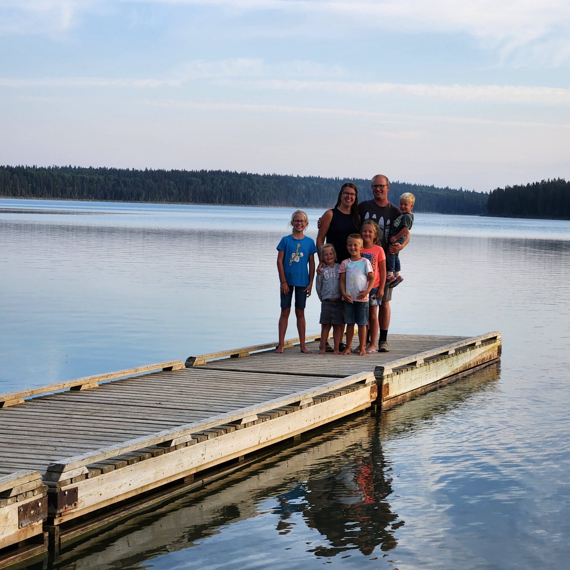 A smiling family of six standing on a long wooden dock over a calm lake under a bright, wispy blue sky.